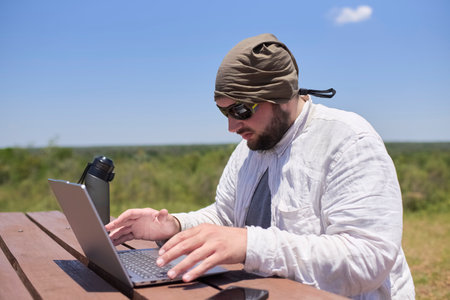 Young Hispanic man working on his laptop at an outdoor table on a sunny day. Concept: digital nomad working remotely while traveling enjoying a lifestyle of freedom.の写真素材