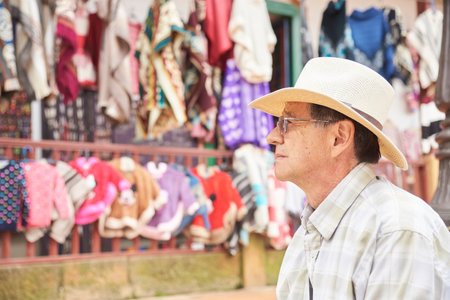 Portrait of a senior Hispanic male seen from the side. It is outdoors and behind it is an artisan clothing store displaying ruanas, ponchos and sweaters of various colors and styles.の写真素材