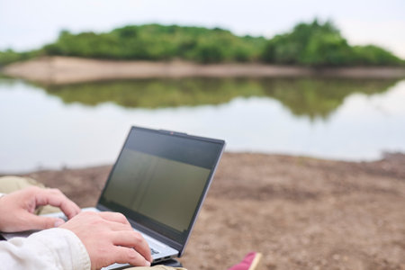 Unknown person sitting outdoors on the bank of a stream, working on his laptop. Concept: digital nomad working remotely while traveling, enjoying a lifestyle of freedom.の写真素材