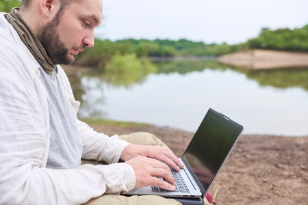 Hispanic man sitting outdoors on the bank of a stream, working on his laptop. Concept: digital nomad working remotely while traveling, enjoying a lifestyle of freedom.の写真素材