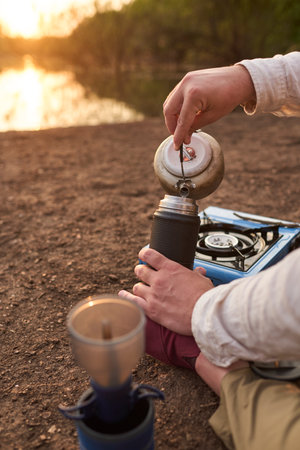 Person pouring hot water from a kettle into a thermos, outdoors, on the bank of a stream, at dawn. Concept: travel to enjoy special moments of tranquility and beauty in nature. Entre Rios, Argentina.の写真素材