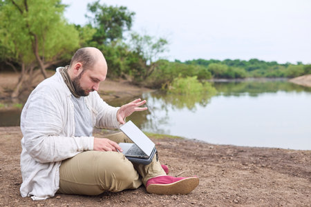 Hispanic man sitting outdoors on the bank of a stream, opening his laptop to work. Concept: digital nomad working remotely while traveling, enjoying a lifestyle of freedom.の写真素材