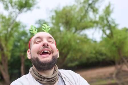A young Hispanic man wearing a funny Christmas reindeer antler headband smiles with a funny expression outdoors, in a natural setting.の写真素材