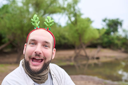 A young Hispanic man wearing a Christmas reindeer antler headband smiles at the camera with a funny expression outdoors, in a natural setting.の写真素材