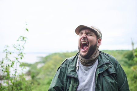 Portrait of a man in nature who appears to be yawning or shouting energetically, with his eyes closed and his mouth wide open.の写真素材