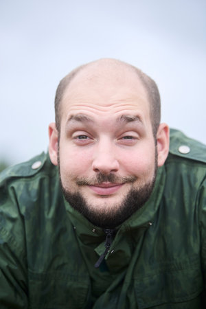 Portrait of a young man looking at the camera with sleepy smile. He is outdoors and his jacket is...