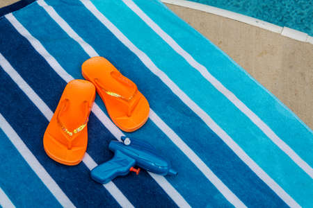 Orange flip flops and squirt gun on beach towel next to poolの写真素材