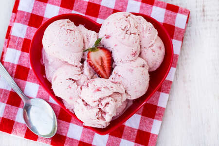 Tasty treat of homemade strawberry ice cream in a red heart shaped bowl with checkered tablecloth and spoonの写真素材