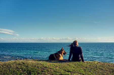 Young woman and her pet German Shepherd dog relaxing overlooking the ocean sitting on a grassy green cliff top looking out over the bay below at sunset in a rear viewの写真素材