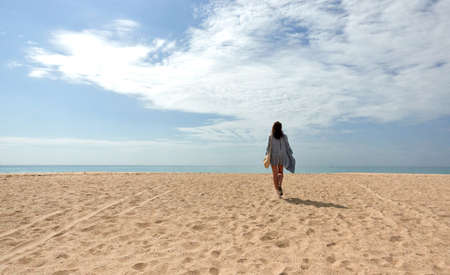 Woman walking away in the distance across a sandy tropical beach to the sea on a sunny spring or summer day with copy space in a vacation or travel conceptの写真素材
