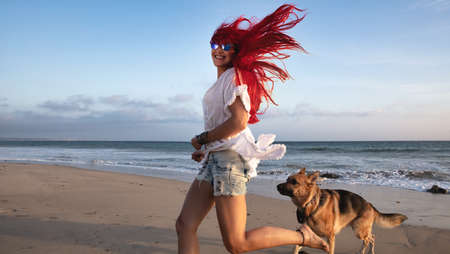 Carefree red-haired young woman running along the beach with a German Shepherd dog in skimpy shorts and sunglasses grinning at the camera with her long red hair flying out behind herの写真素材