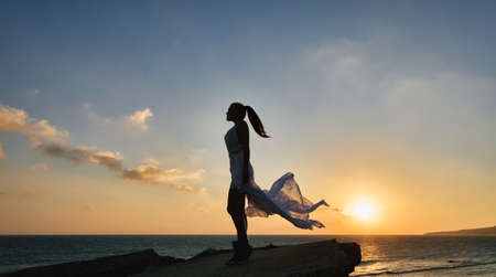 Trendy and sexy beautiful woman standing on a rock at the seaside in a white swimsuit and flowing sarong silhouetted against the colorful orange tropical sunset reflected on the oceanの写真素材