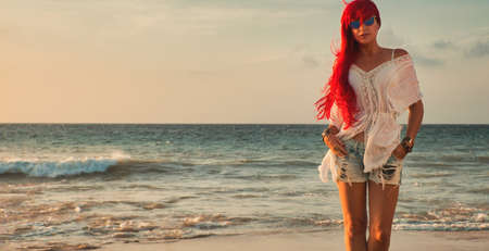 Beautiful red haired young woman in casual summer style on the beach at sunsetの写真素材