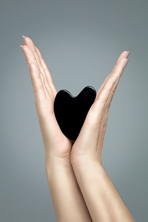 Female hands with an obsidian guasha on a gray background. Anti aging beauty and skincare concept. Vertical format with copy spaceの写真素材