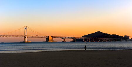 A lonely jogger on a beach on sunset with an impressive suspended bridge in backgroundの写真素材