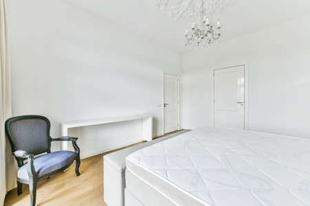 Comfortable armchair and empty shelf placed near bed with soft mattress against white walls with...