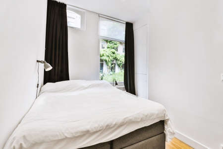 Comfortable bed and lamp placed in small narrow minimalist style bedroom with white walls and black...