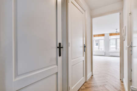 Narrow corridor with white walls and doors leading to spacious room with windows and parquet floor in modern apartmentの写真素材