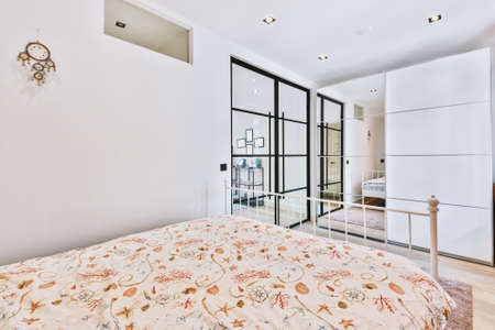Comfortable bed with ornamental blanket placed near glass door and wardrobe with mirror in light...