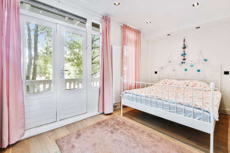 Comfortable bed located near balcony door with curtains in cozy light bedroom at homeの写真素材
