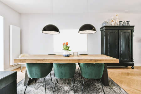 Comfortable chairs and wooden table with flowers placed under stylish lamps against wall with painting and cupboard in dining roomの写真素材