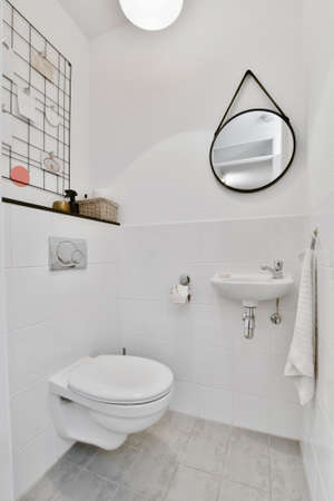 Modern flush toilet and ceramic sink installed on white tiled walls near mirror and towel in small...