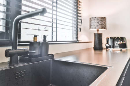 Shiny counter of kitchen with black washbasin and faucet near window with black blinds