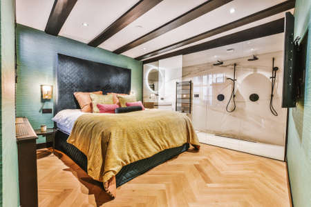 Interior of stylish bedroom with comfortable bed located near glass wall of bathroom in hotel