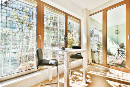 Sun lit room with small table and stools near panoramic window viewing modern city buildings