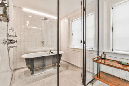 Glass wall in modern white bathroom with black soaker tub and small wooden table at window