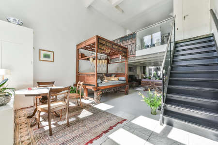 Sun lit room with open second floor and black staircase and ornamental wooden furniture in house