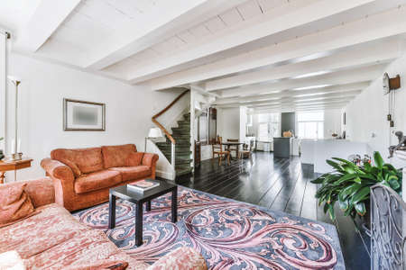 House sun lit room with terracotta sofas and ornamental carpet on wooden floor near staircase...