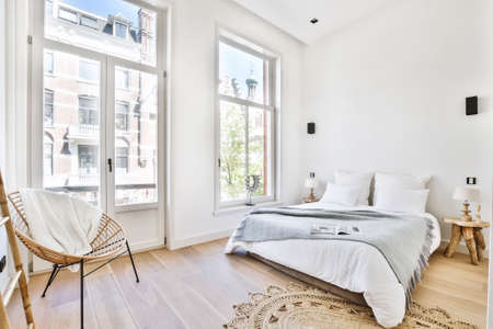 Comfortable bed with blanket and pillows located near lamps and chair in spacious bedroom in modern apartmentの写真素材