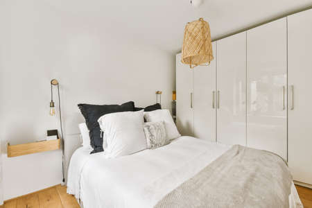 Comfortable bed with wardobe, bedside table with lamp placed near window in light bedroom at home