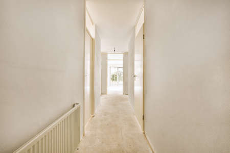 Narrow corridor with white walls and doors leading to spacious room with windows and parquet floor...