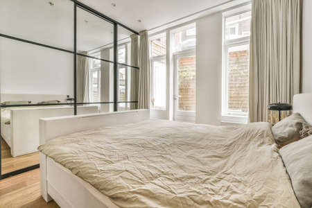 Comfortable bed with white bedclothes and bedside table with lamp placed near window and balcony...
