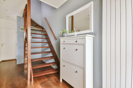 White chest of drawers and a mirror next to a brown wooden staircase connecting the levels of a...
