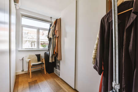 Dressing room with window, chair, white cabinets and hangers with clothes
