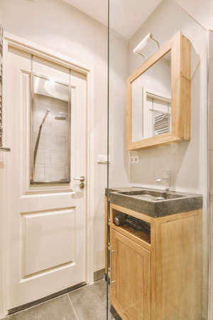 Cupboard with sink and mirror attached to tiled wall near door and drying rack in modern restroomの写真素材