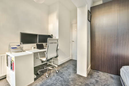 Comfortable chair placed near desk with computer monitors and whiteboard in light workplace in...