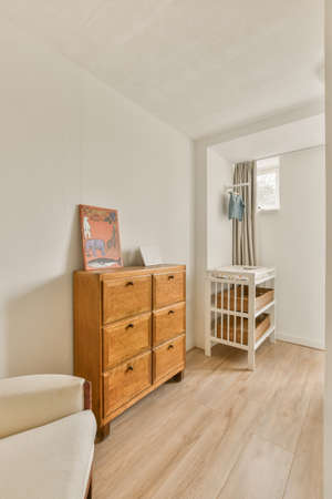 Interior of white room with baby crib and armchair near window with blue chest of drawers in...
