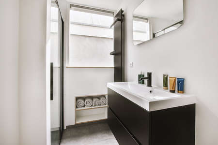 Cupboard with sink and mirror attached to tiled wall near door and drying rack in modern restroomの写真素材