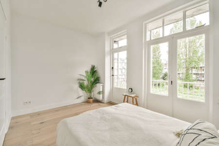 Comfortable bed with white bedclothes and bedside table with lamp placed near window and balcony...