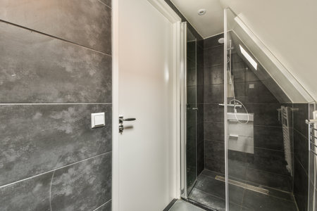 Tiled shower cabin with glass door located near small window in mansard apartment