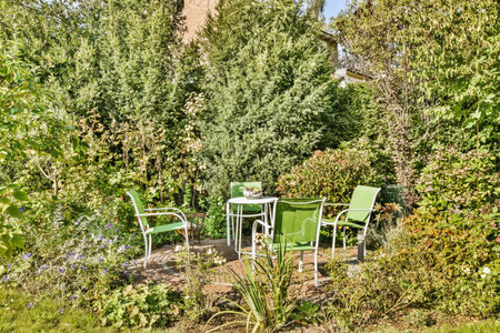 two green chairs in the middle of a garden, surrounded by trees and bushes with blue sky in the...