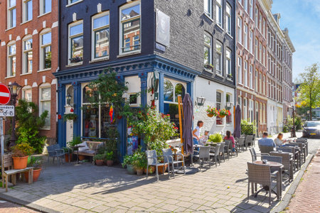Amsterdam, Netherlands - 10 April, 2021: an outdoor cafe with tables and chairs on the sidewalk in front of a row of tall brick buildings, surrounded by treesのeditorial素材