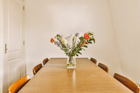 some flowers in a vase on a dining room table with white walls and light wood chairs around the wooden tableの写真素材