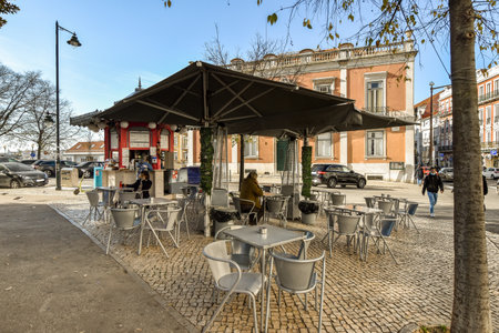 some tables and chairs under an umbrella on a sidewalk in front of a building with people walking by the streetのeditorial素材