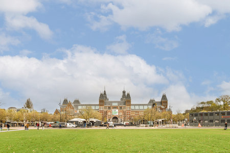 a city park with people walking and sitting on the green grass in front of some buildings under a cloudy blue skyのeditorial素材
