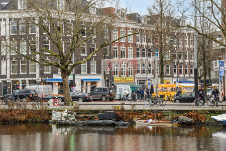 Amsterdam, Netherlands - 10 April, 2021: some boats in the water and buildings on the other side of the river, with people walking along the shoreのeditorial素材